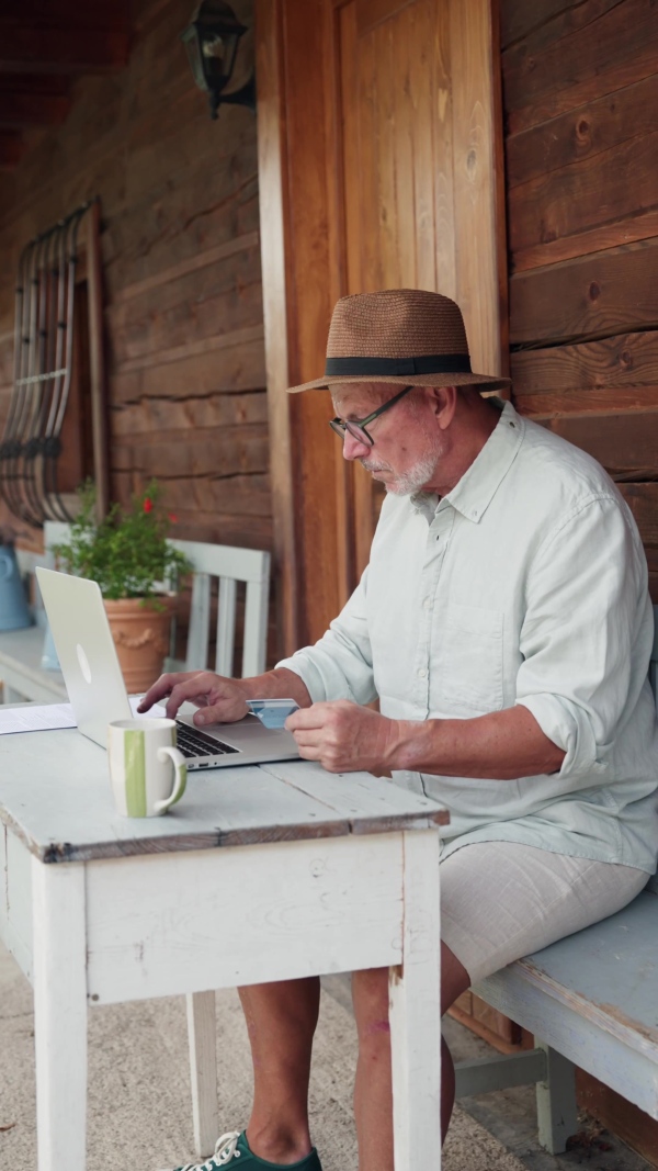 A senior man sitting outdoor on terrace, shopping online on notebook. Man holding debit card in hand, imputing card detail for payment.