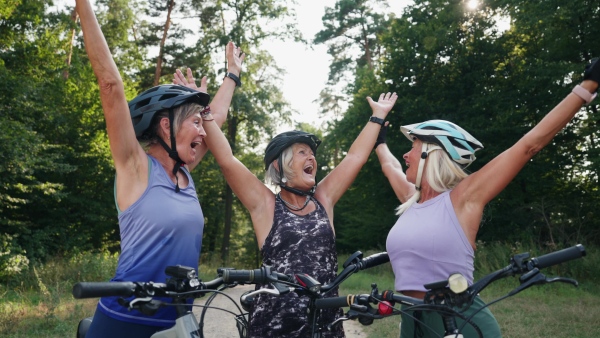 Three senior friends on their bike tour in nature resting, celebrating successful ride. Older women on low impact route riding electric bikes.