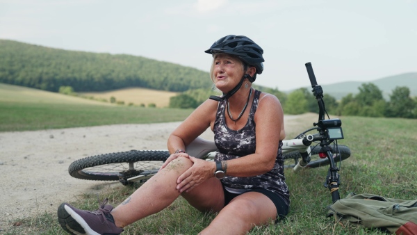 Female senior cyclist fell off her bike, injuring her knee. Elderly woman in pain sitting on ground, holding her leg and crying.