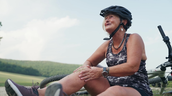 Female senior cyclist fell off her bike, injuring her knee. Elderly woman in pain sitting on ground, holding her leg and crying.