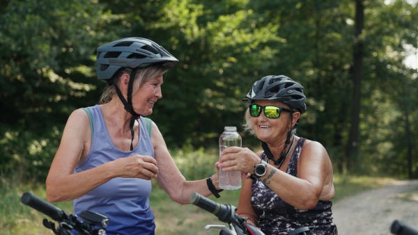 Senior friends on their bike tour in nature resting, drinking water. Older women on low impact route riding electric bikes.