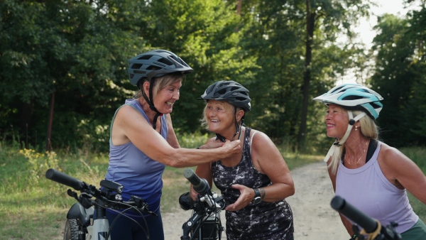 Three senior friends preparing for their bike tour in nature, putting helmets on. Older women on low impact route riding electric bikes.