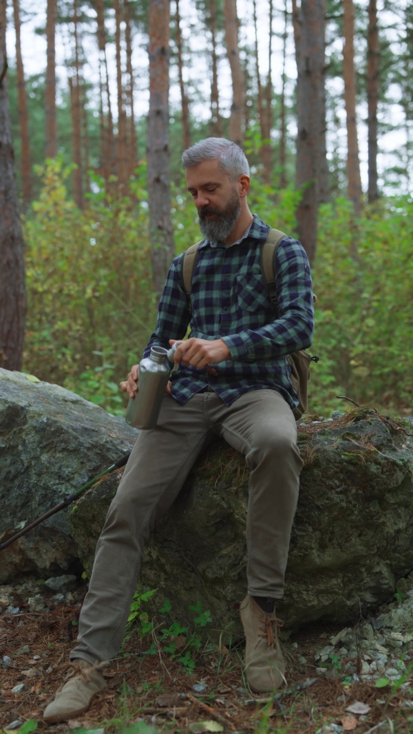 Handsome mature man sitting on rock in the middle of nature, enjoying peaceful atmosphere of the forest, drinking water.