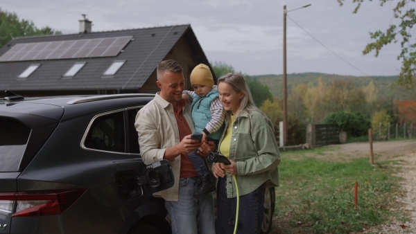 Parents with their son charging their electric car in front of house. Checking charging progress on smartphone.