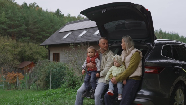 A family sitting in trunk of electric car parked in front of house. House with rooftop solar panels and own EV home charging station.