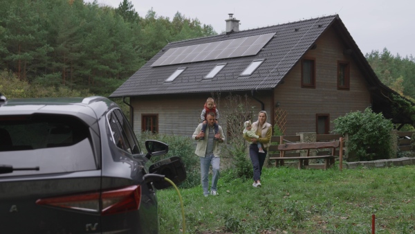 Parents with two kids walking to their electric car parked in front of house. House with rooftop solar panels and own EV home charging station.