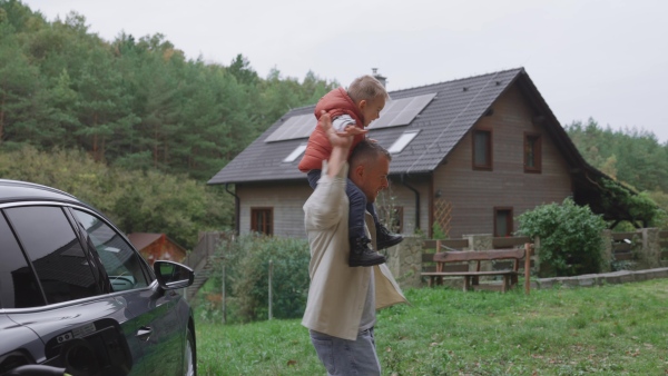 Father playing with his son carry him on shoulders by electric car and in front of house with solar panels on rooftop.