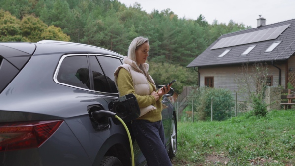 Young woman holding smartphone, using app for charging her electric car. EV home charging station.