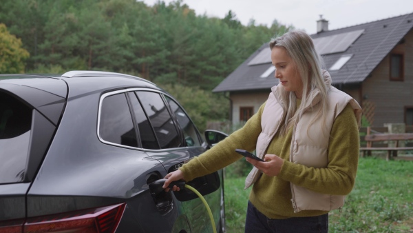 Young woman holding smartphone, using app for charging her electric car. EV home charging station.