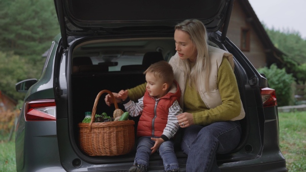 Mom and little boy sitting in trunk of electric car parked in front of house. House with rooftop solar panels and own EV home charging station.