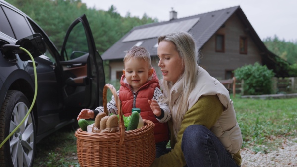 A mother and son after grocery shopping by electric car parked in front of house. House with rooftop solar panels and own EV home charging station.