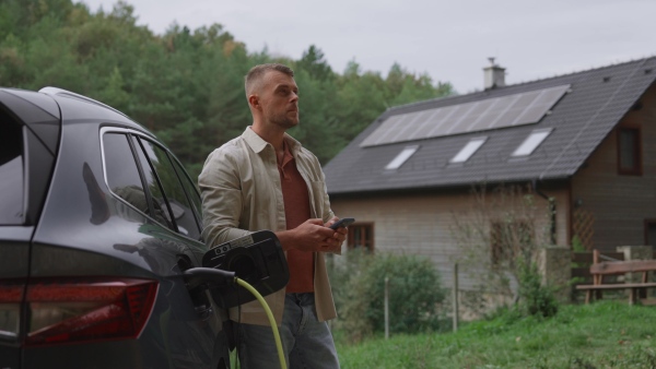 A handsome man leaning against electric car holding smartphone in hand. Electric vehicle with charger in charging port.