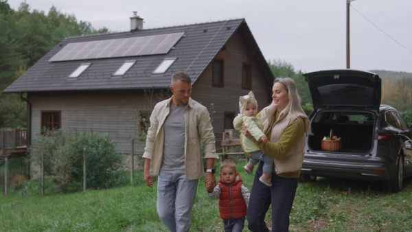 A family in front electric car with open trunk. House with rooftop solar panels and own EV home charging station.