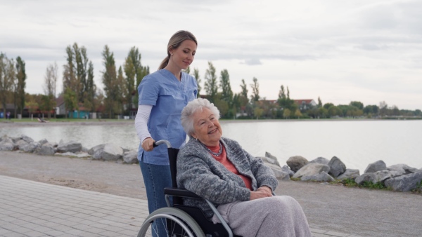 Disabled senior woman and her home caregiver spending a chilly, windy day outdoors by lake. Autumn walk for elderly patient in wheelchair.