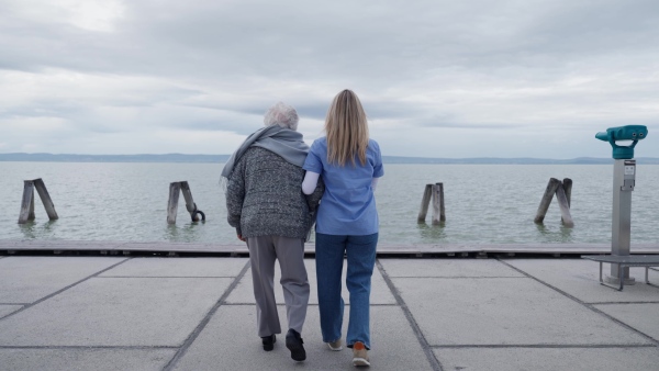 A rear view of senior woman and her home caregiver spending a chilly, windy day outdoors by lake. Autumn walk for elderly patient.