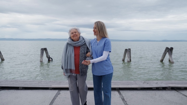 A senior woman and her home caregiver spending a chilly, windy day outdoors by lake. Autumn walk for elderly patient.