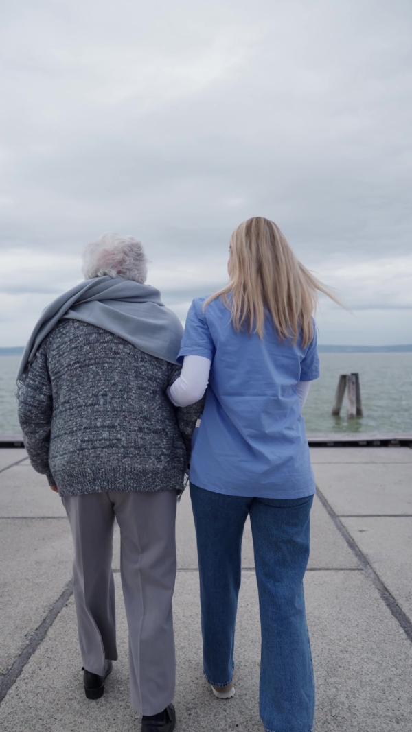 A rear view of senior woman and her home caregiver spending a chilly, windy day outdoors by lake. Autumn walk for elderly patient.