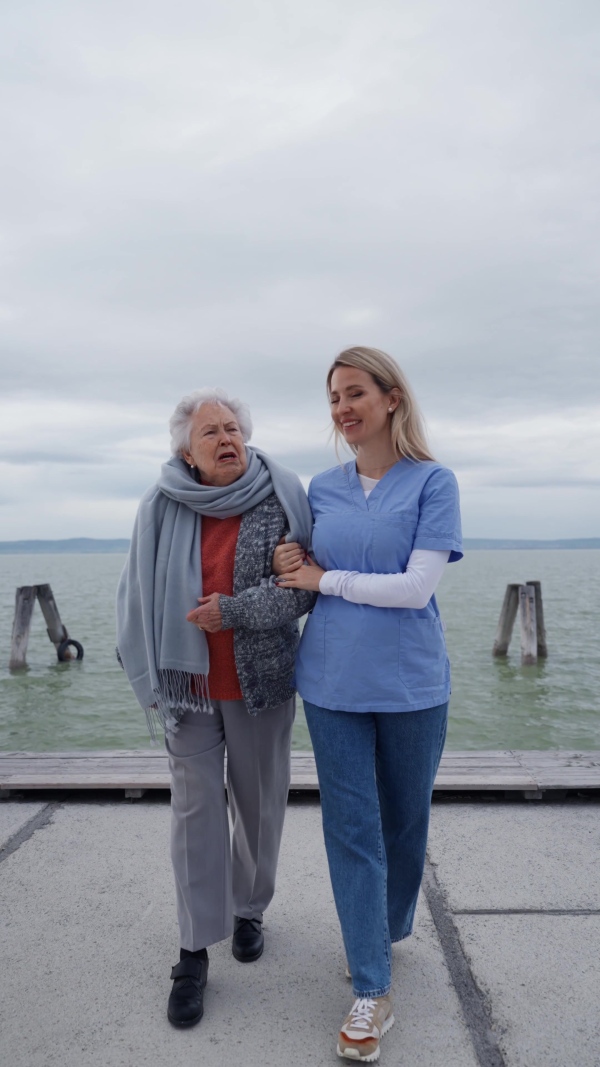 A senior woman and her home caregiver spending a chilly, windy day outdoors by lake. Autumn walk for elderly patient.