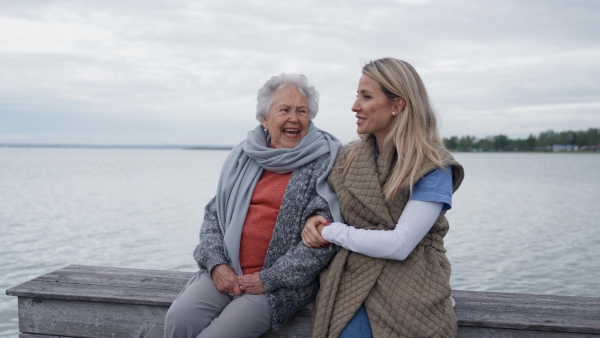 Senior woman and her home caregiver spending a chilly day outdoors, sitting by lake. Autumn walk for elderly patient.