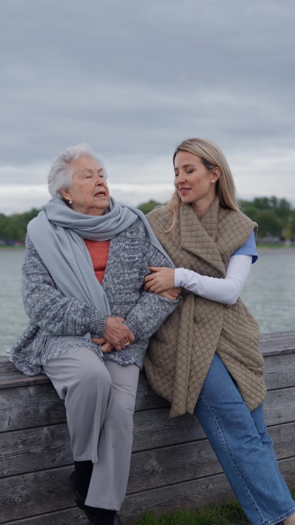 Senior woman and her home caregiver spending a chilly day outdoors, sitting by lake. Autumn walk for elderly patient.