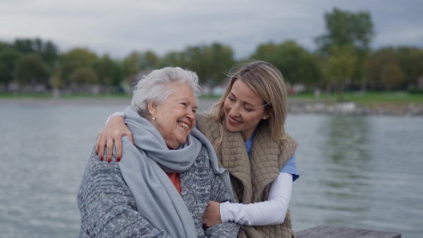 Senior woman and her home caregiver spending a chilly day outdoors, sitting by lake. Autumn walk for elderly patient.