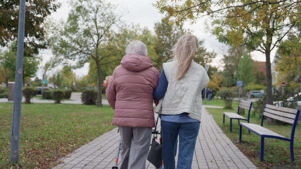 Rear view of senior woman and her home caregiver spending a chilly, windy day outdoors in city park. Autumn walk for elderly patient with walker.