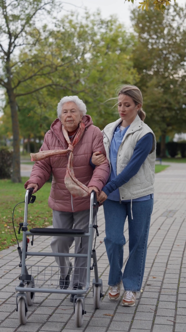 Senior woman and her home caregiver spending a chilly, windy day outdoors in city park. Autumn walk for elderly patient with walker.