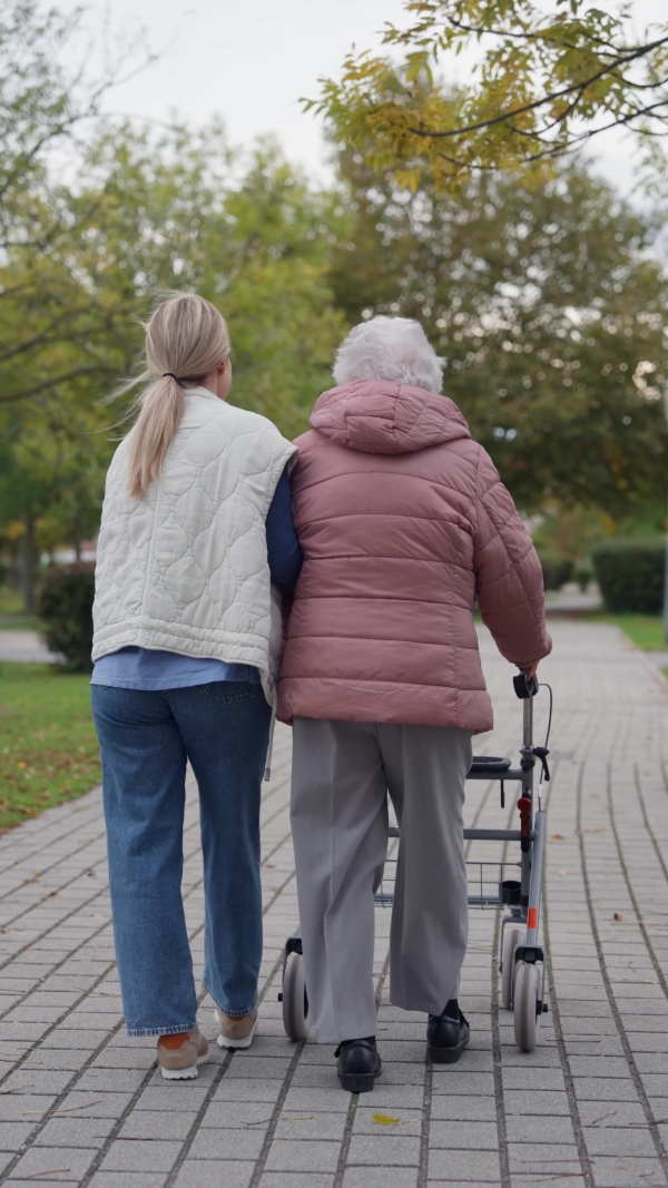 Rear view of senior woman and her home caregiver spending a chilly, windy day outdoors in city park. Autumn walk for elderly patient with walker.