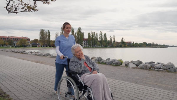 Disabled senior woman and her home caregiver spending a chilly, windy day outdoors by lake. Autumn walk for elderly patient in wheelchair.