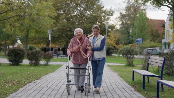 Senior woman and her home caregiver spending a chilly, windy day outdoors in city park. Autumn walk for elderly patient with walker.