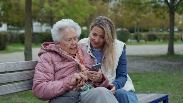 A nurse and female senior patient sitting on bench in the park, looking at family photos on mobile phone.