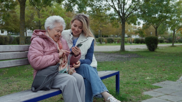 A nurse and female senior patient sitting on bench in the park, looking at family photos on mobile phone.