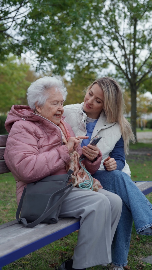 A nurse and female senior patient sitting on bench in the park, looking at family photos on mobile phone.