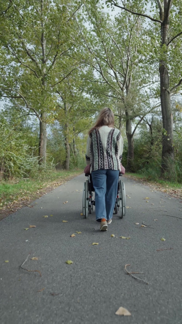 An autumn walk with wheelchair, disabled senior woman and her daughter spending a chilly, windy day outdoors in nature. Rear view.