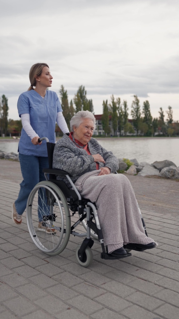 Disabled senior woman and her home caregiver spending a chilly, windy day outdoors by lake. Autumn walk for elderly patient in wheelchair.