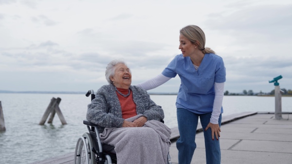 Disabled senior woman and her home caregiver spending a chilly, windy day outdoors by lake, looking at water. Autumn walk for elderly patient in wheelchair.