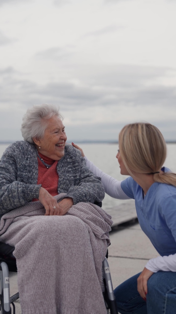 Disabled senior woman and her home caregiver spending a chilly, windy day outdoors by lake, looking at water. Autumn walk for elderly patient in wheelchair.