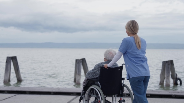 Disabled senior woman and her home caregiver spending a chilly, windy day outdoors by lake, looking at water. Autumn walk for elderly patient in wheelchair.