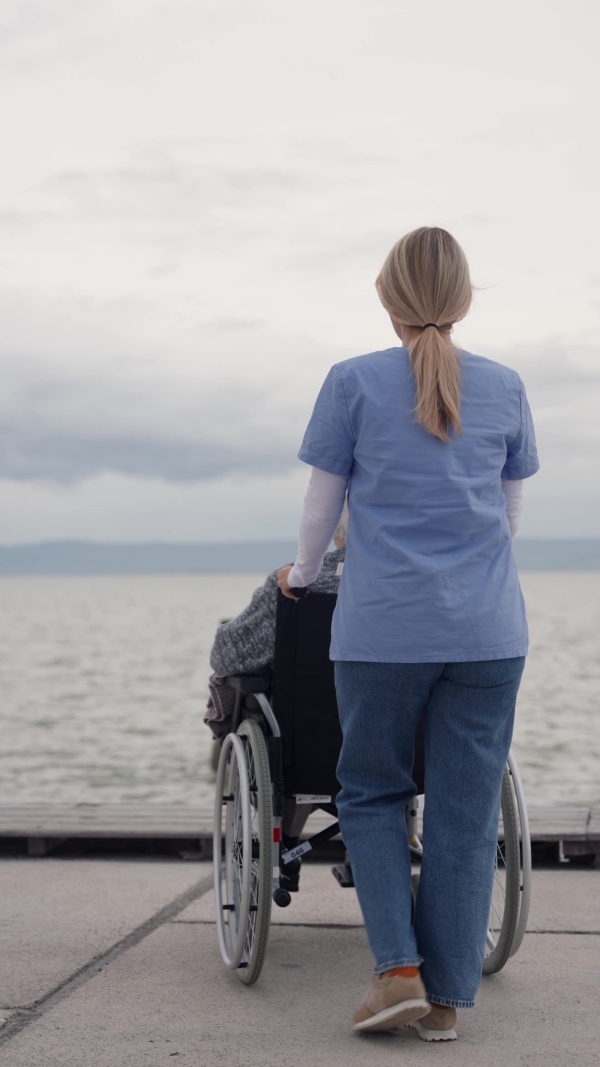 Disabled senior woman and her home caregiver spending a chilly, windy day outdoors by lake, looking at water. Autumn walk for elderly patient in wheelchair.