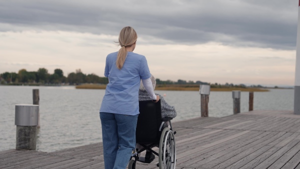 Disabled senior woman and her home caregiver spending a chilly, windy day outdoors by lake, looking at water. Autumn walk for elderly patient in wheelchair.