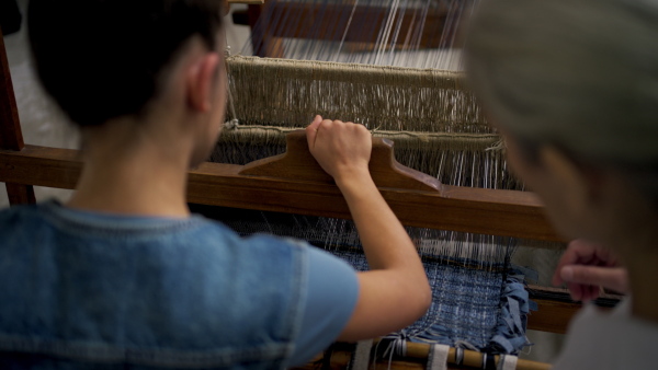 A rear view of mother and daughter working on wooden loom, weaving. Sustainable business, using recycled materials in fashion.