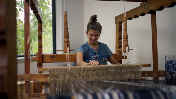 A teenage girl weaving on wooden loom, working in family weaving workshop. Sustainable business, using recycled materials in fashion.