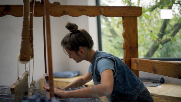 A teenage girl weaving on wooden loom, working in family weaving workshop. Sustainable business, using recycled materials in fashion.