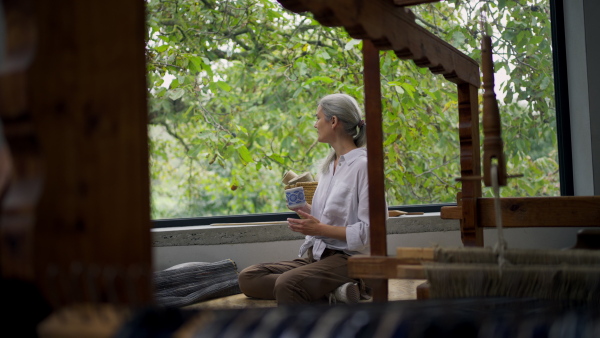 A side view of woman sitting by window, holding cup of coffee and taking a break.