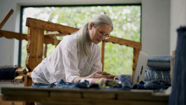 A woman preparing fabrics for new product in weaving workshop, checking customer order on laptop. Sustainable business.