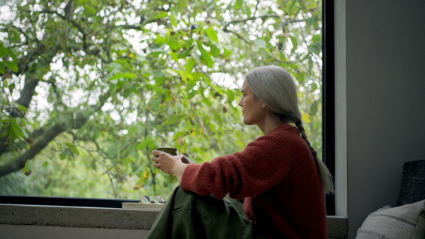 A side view of woman sitting by window, holding cup of coffee and taking a break.