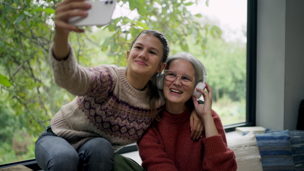 Mom and her daughter sitting by big window, taking selfie.