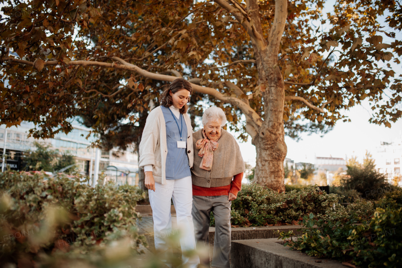 Portrait of nurse and elderly woman on walk in park during warm autumn day. Young caregiver spending time with senior patient.