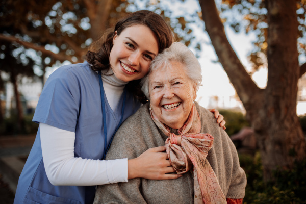 Portrait of nurse and elderly woman on walk in park during warm autumn day. Young caregiver spending time with senior patient.