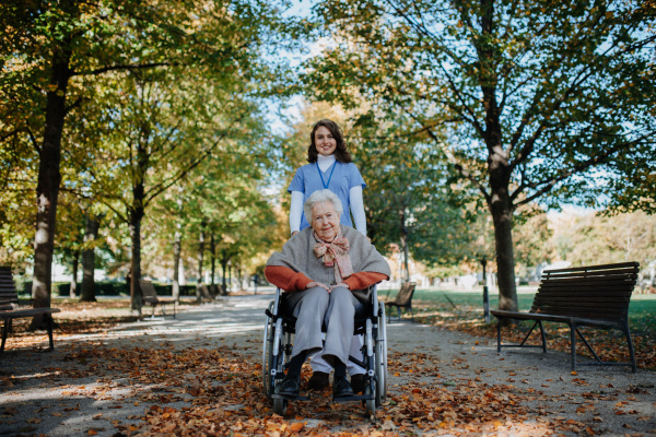 Disabled elderly woman in wheelchair on walk in park during warm autumn day. Young nurse pushing wheelchair, talking with senior woman.